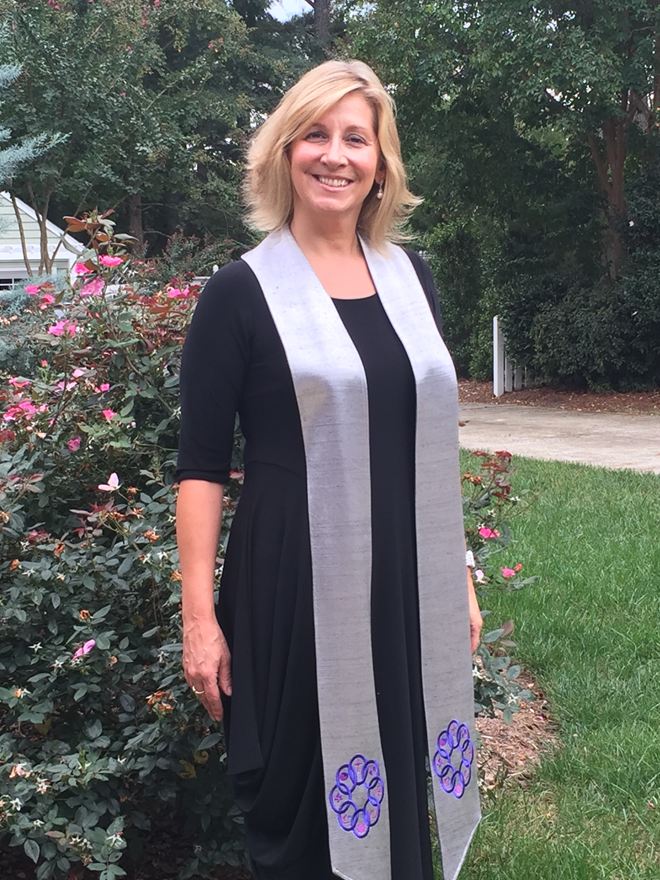 Rev. Christie Mabry smiling in a garden beside pink roses, in a black dress and a silver stole embroidered with purple knotwork