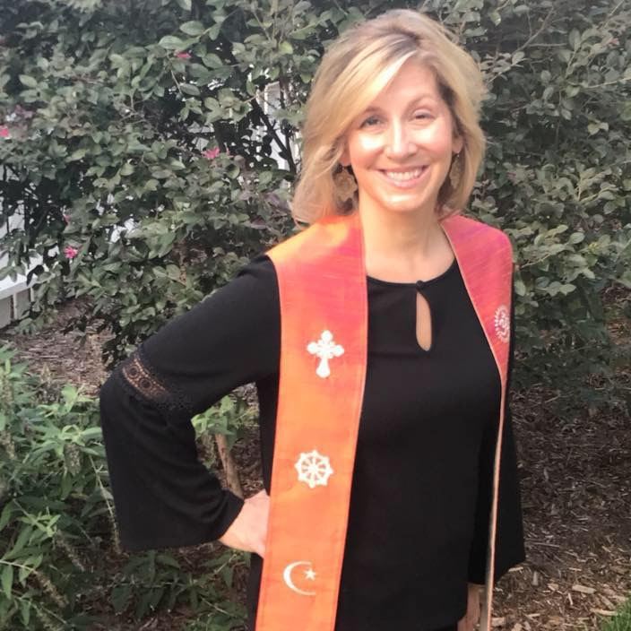 Rev. Christie Mabry smiling outdoors among green shrubs, one hand on her hip, in her orange stole embroidered with a cross, a dharma wheel and a crescent moon