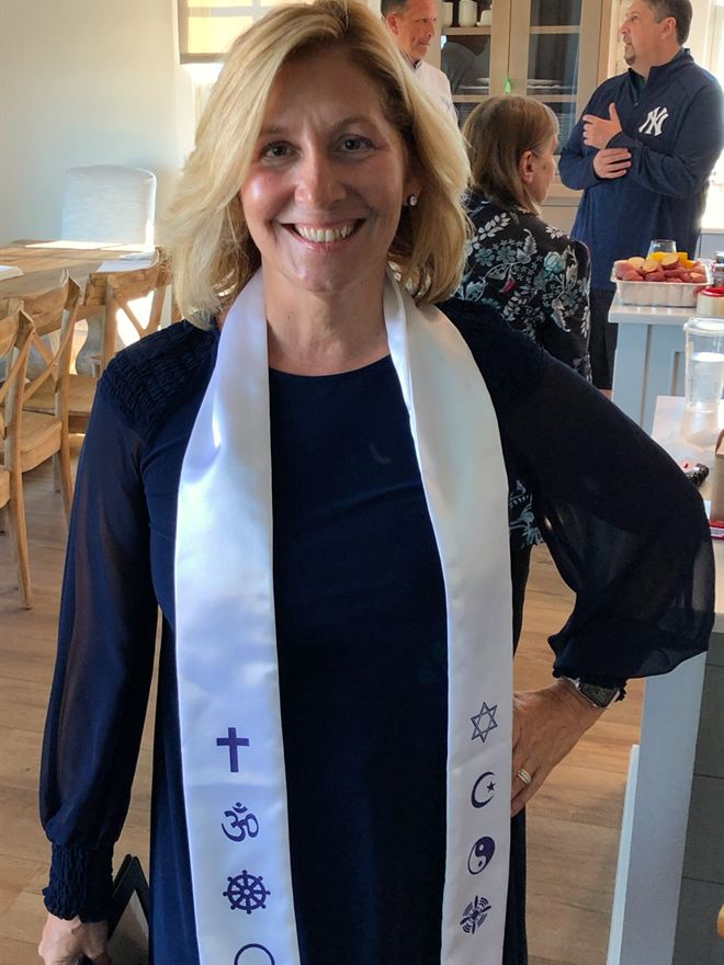 Rev. Christie smiling in a white stole printed with a cross, an om, a dharma wheel, a Star of David, a crescent moon and a yin-yang
