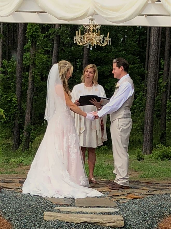 Rev. Christie reading from her ceremony book as a bride and groom hold hands beneath a draped pergola in the pines