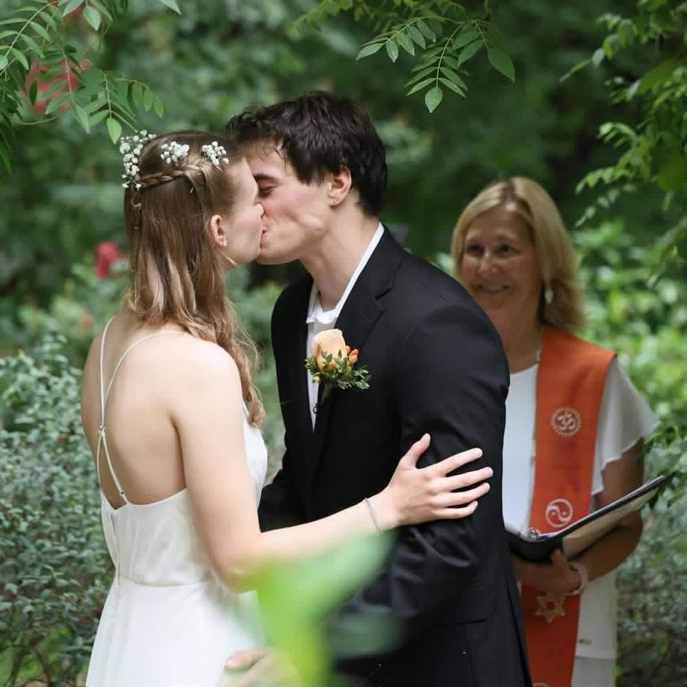 A couple shares their first kiss in a leafy garden while Rev. Christie Mabry smiles beside them in her interfaith stole
