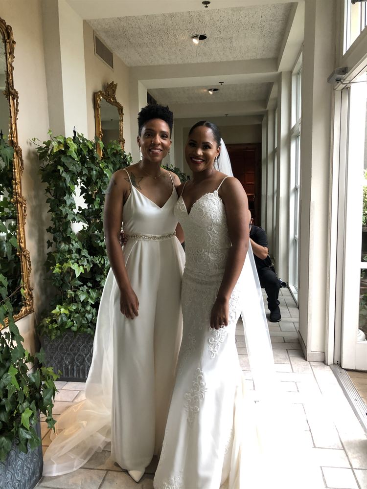 Two brides smiling arm in arm in a sunlit conservatory filled with ivy