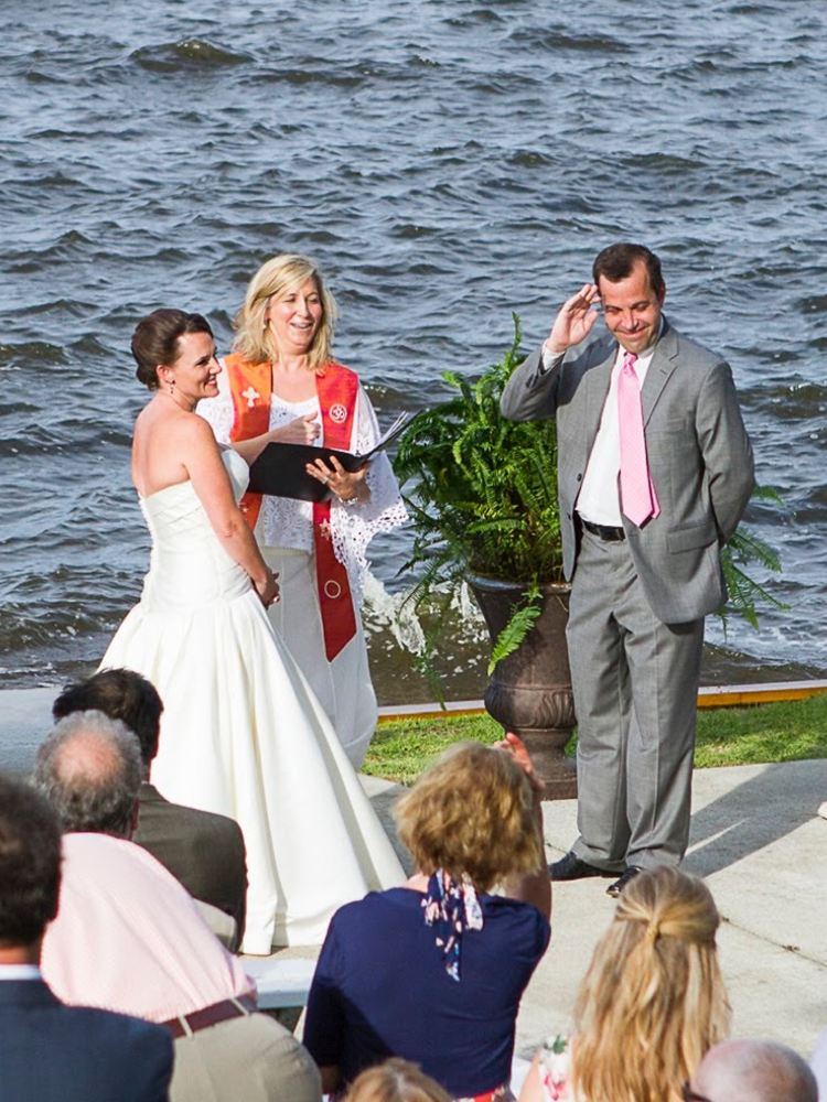 Rev. Christie, in her orange interfaith stole, laughing with a bride and groom at a waterfront ceremony