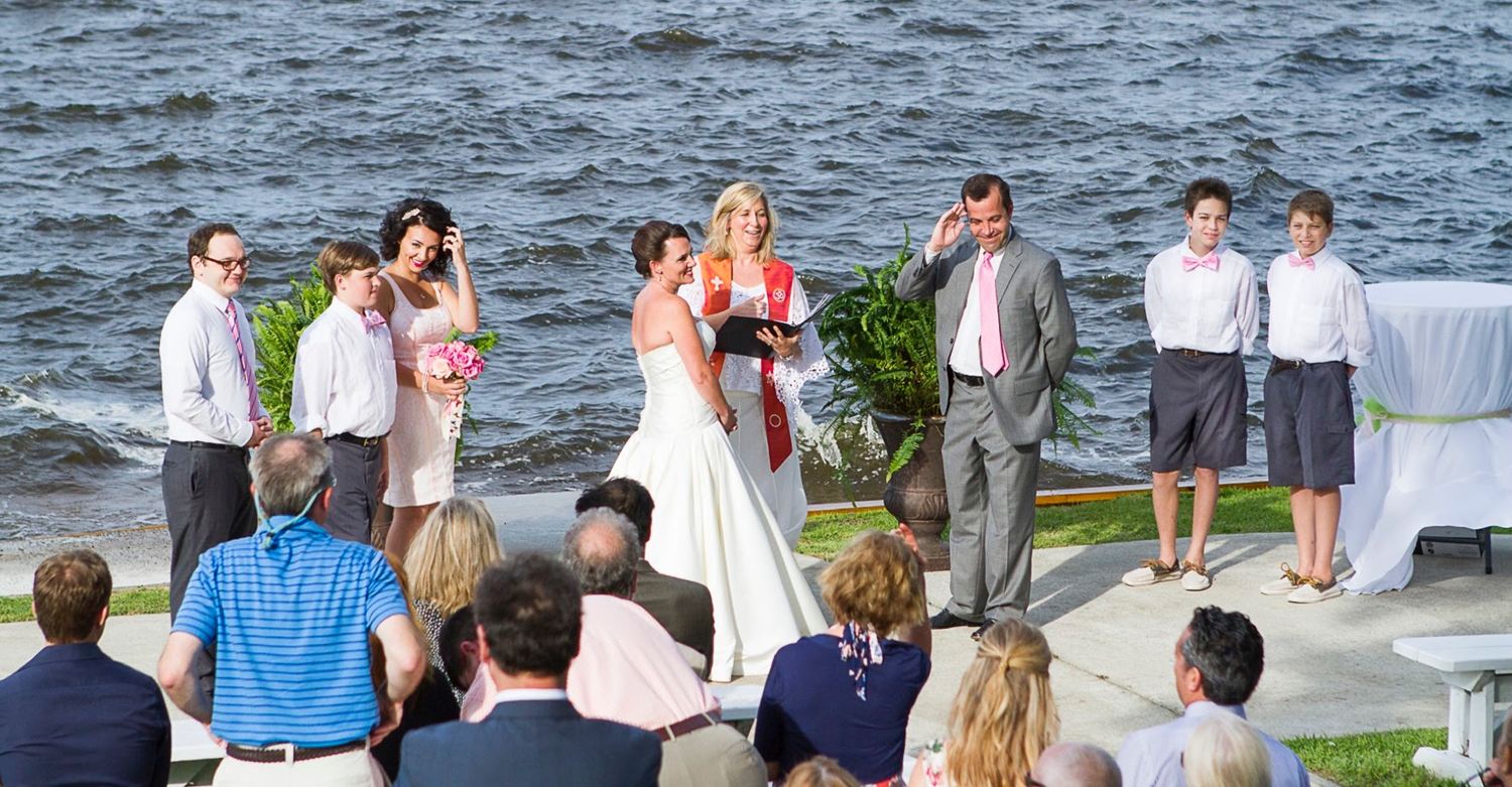 Rev. Christie, in her orange interfaith stole, officiating a waterfront ceremony as the bride and groom laugh and their guests look on
